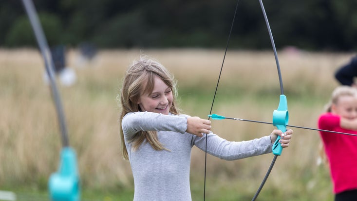 A girl in a grey top smiling while pulling back an archery bow and arrow